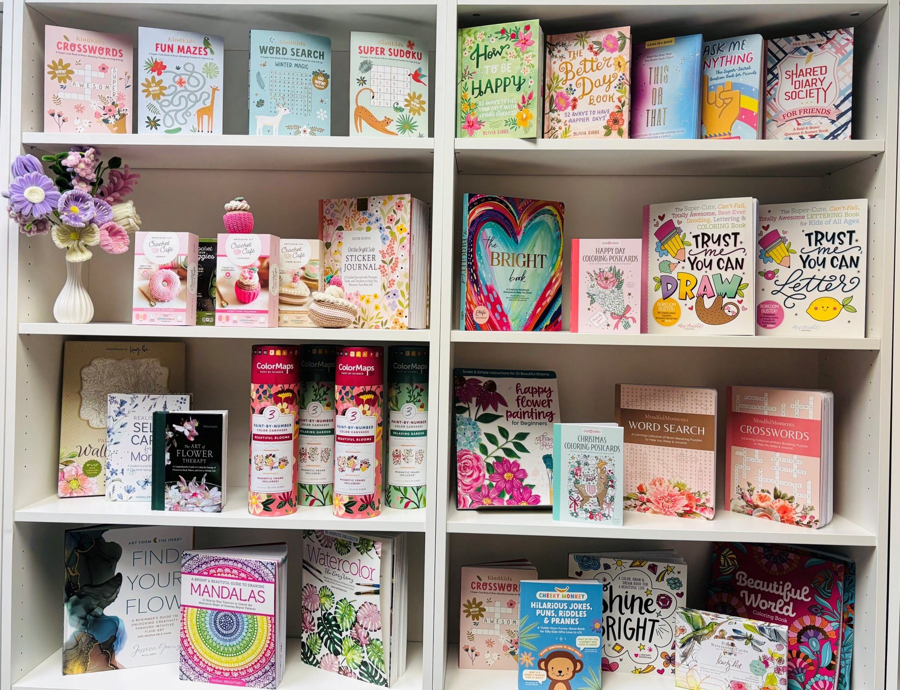 A white two-bay bookcase of colorful Schiffer Publishing activity books with a small floral arrangement