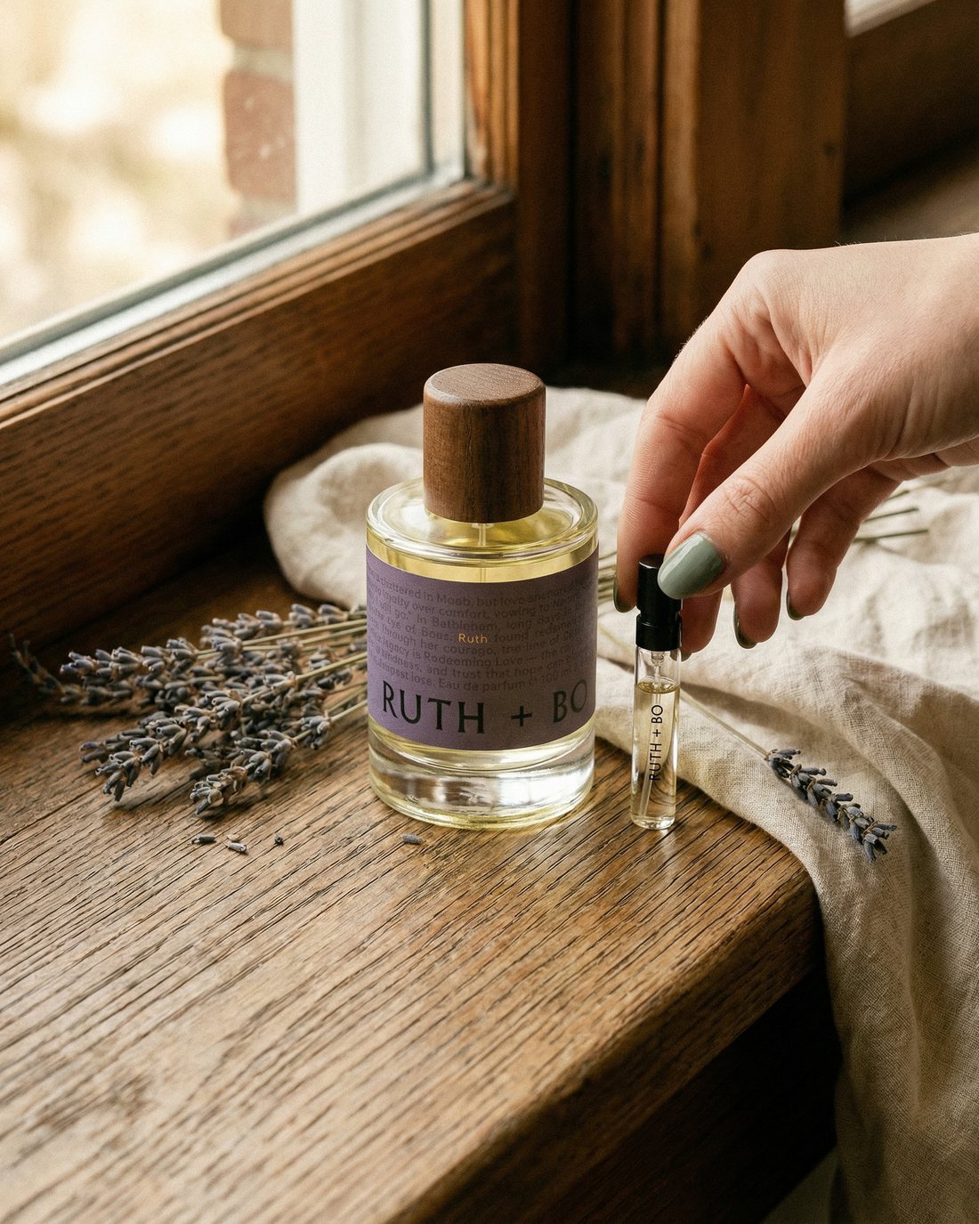 A hand lifting a Ruth + Bo tester beside the perfume bottle on a sunny windowsill